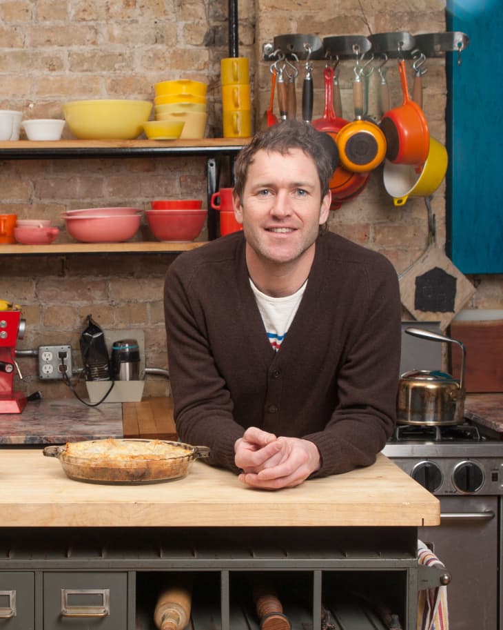 Man in a kitchen with brick walls, leaning on a wooden counter with a baked dish, colorful cookware on shelves.