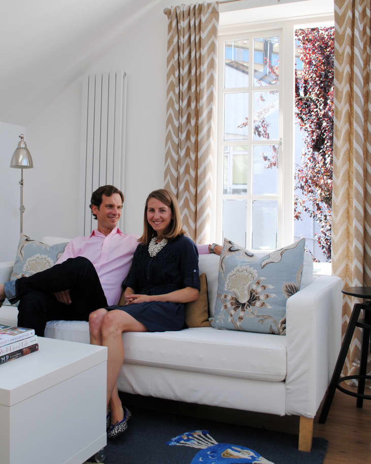 Couple sitting on a white sofa with floral pillows, next to a window with chevron curtains and a peacock rug on the floor.