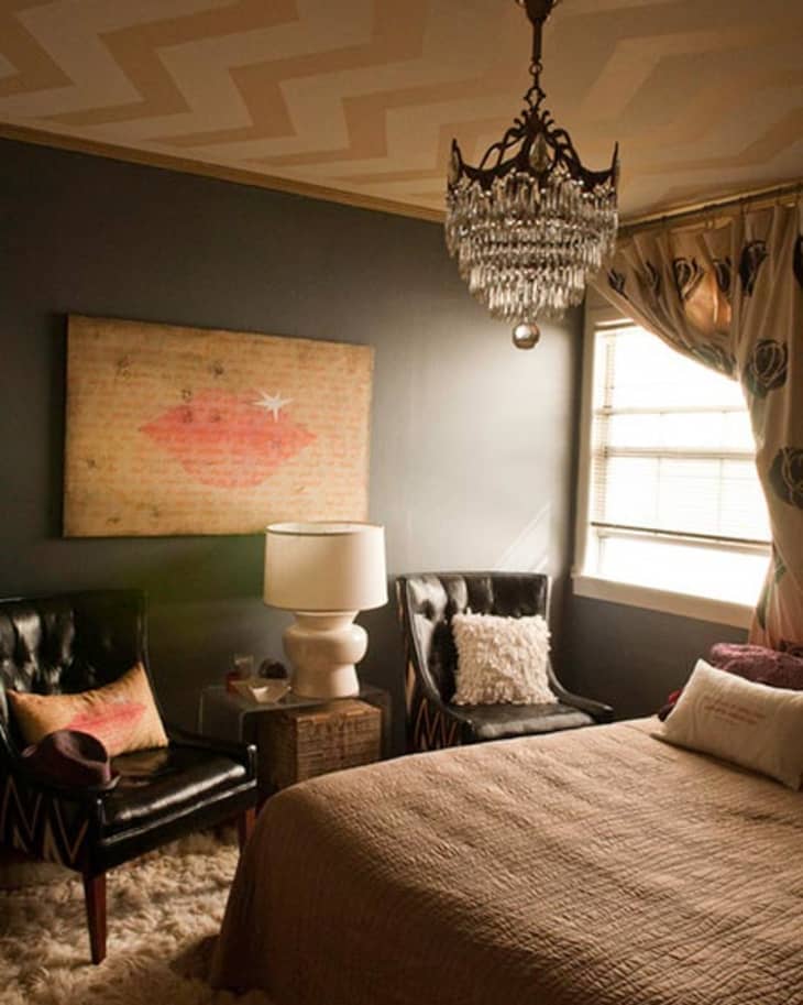 Cozy bedroom with dark walls, chandelier, two black chairs, a lamp, and a bed with brown bedding.