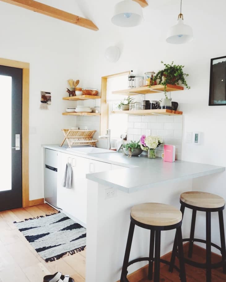 Modern kitchen with white cabinets, open shelves, plants, and wooden stools at a breakfast bar.