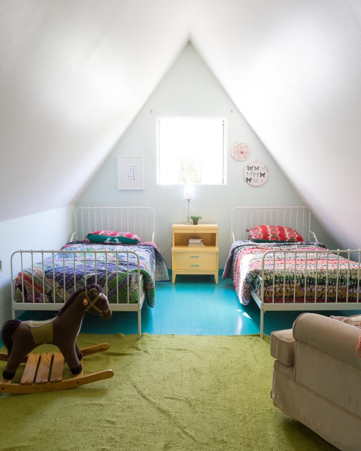 Attic bedroom with two metal beds, colorful quilts, a wooden rocking horse, and a green rug.