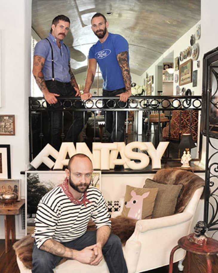 Three men in a stylish room with eclectic decor, including a "FANTASY" sign and various framed artworks.
