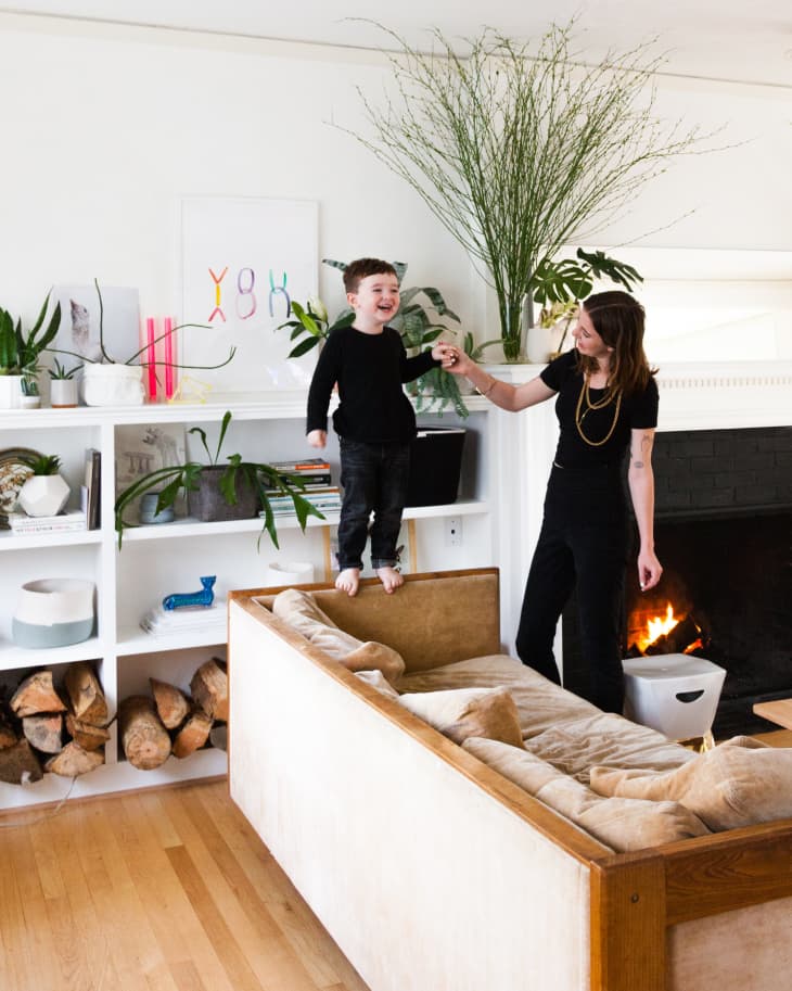 Child standing on a beige sofa, holding hands with a woman in a living room with plants and a fireplace.