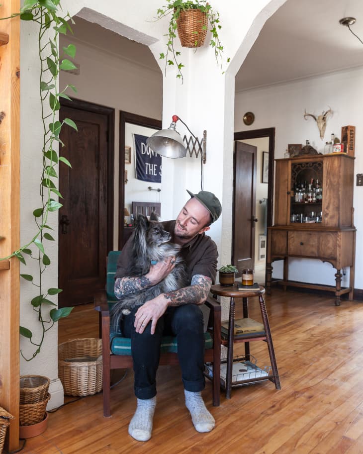 Man with tattoos sitting on a chair, holding a dog in a cozy living room with plants and wooden furniture.