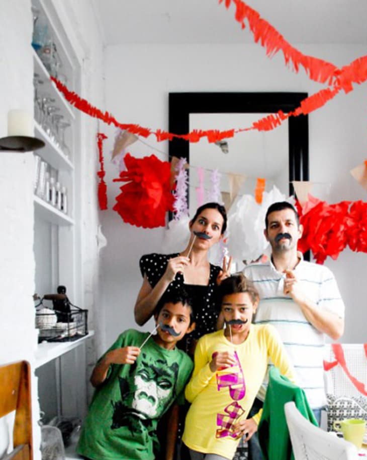 Family posing with fake mustaches in a decorated room with red and white paper garlands.