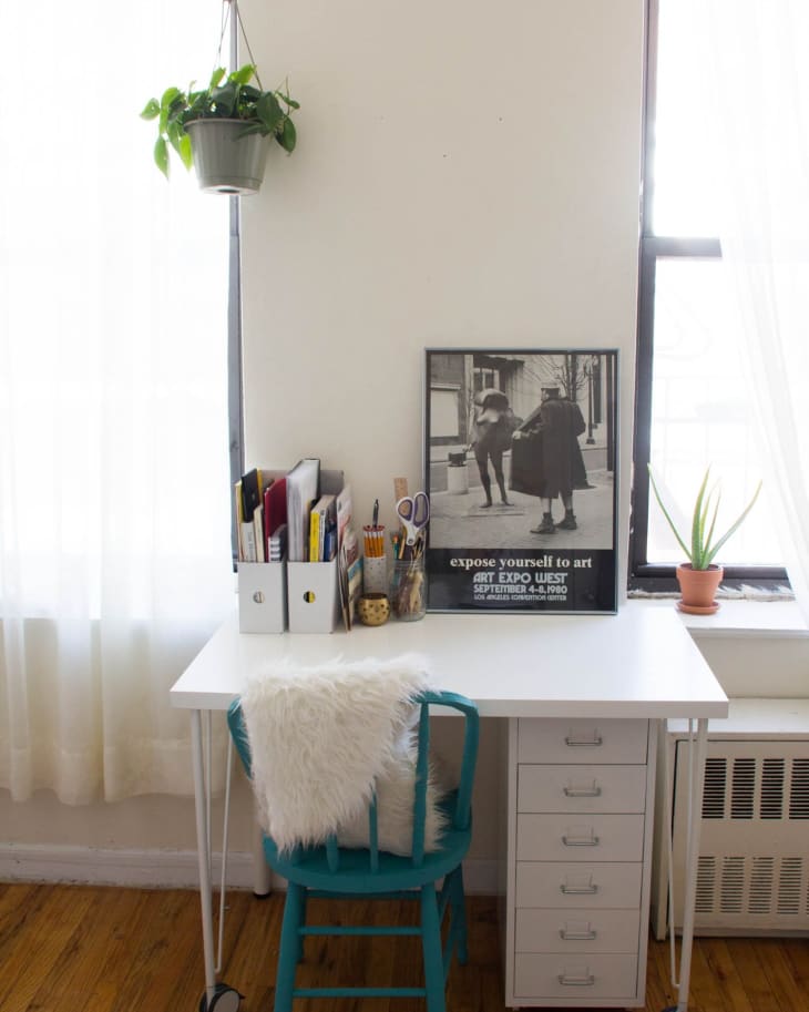 White desk with teal chair, hanging plant, art poster, and stationery organizer by a window.