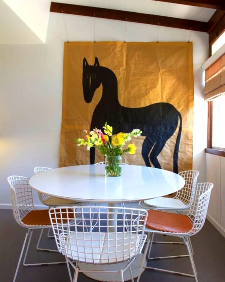 Dining area with a round white table, wire chairs, floral centerpiece, and large horse artwork on the wall.