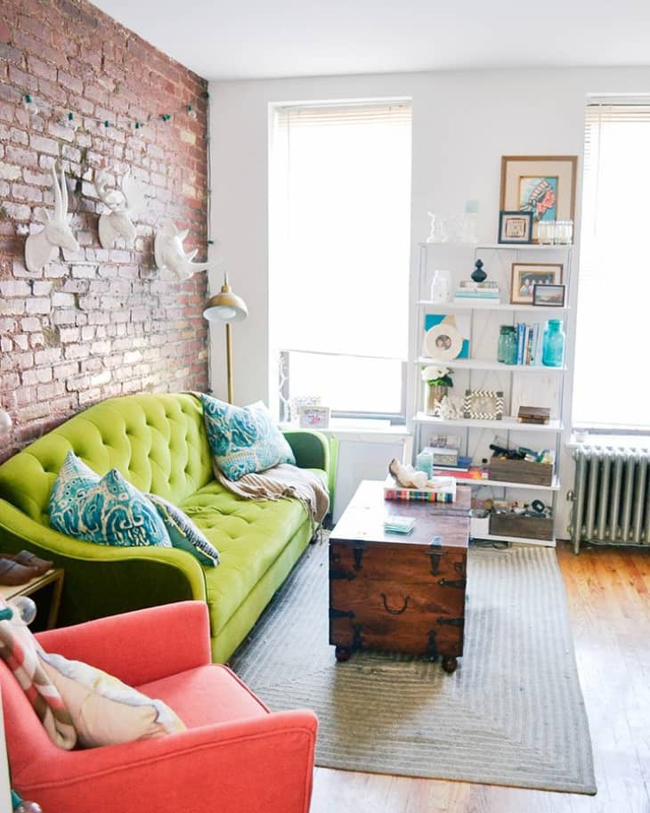 Living room with green sofa, coral armchair, wooden chest coffee table, brick wall, and white shelving unit.