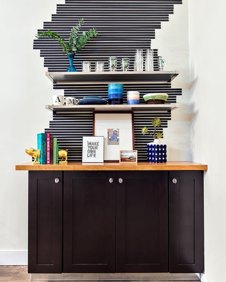 Black cabinet with wooden top, decorative items, books, and glassware on shelves against a striped wall.