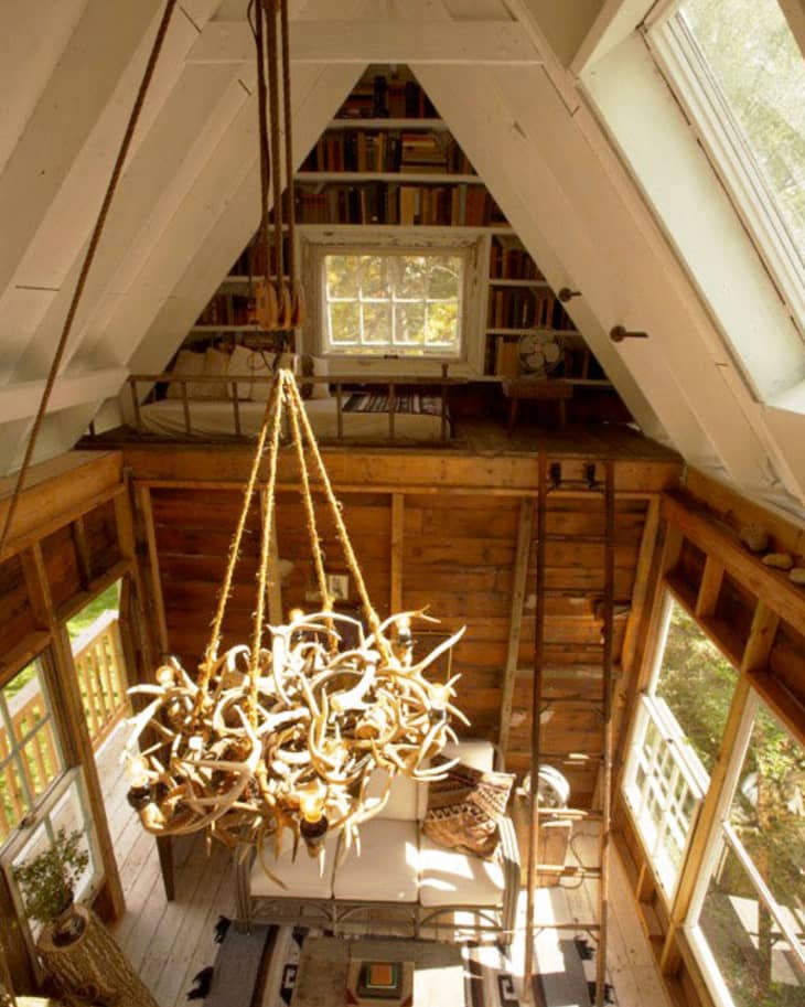 A-frame cabin interior with antler chandelier, loft library, and large windows.