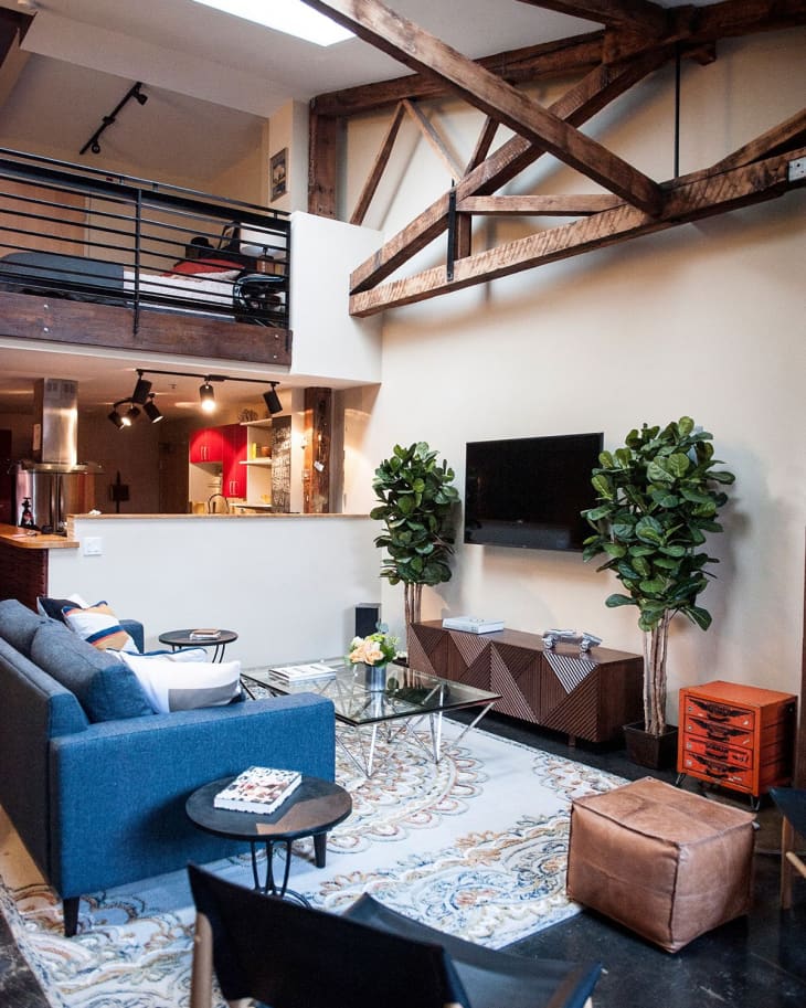 Loft living room with blue sofa, glass coffee table, patterned rug, TV, and exposed wooden beams.