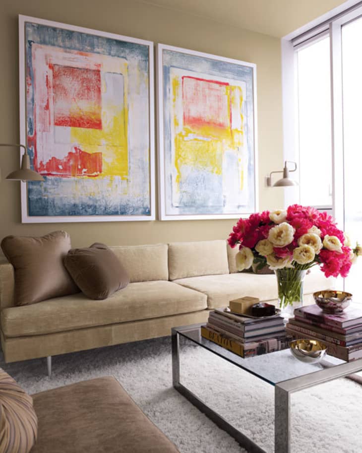 Living room with beige sofa, abstract art, glass coffee table, and pink and white flowers in a vase.