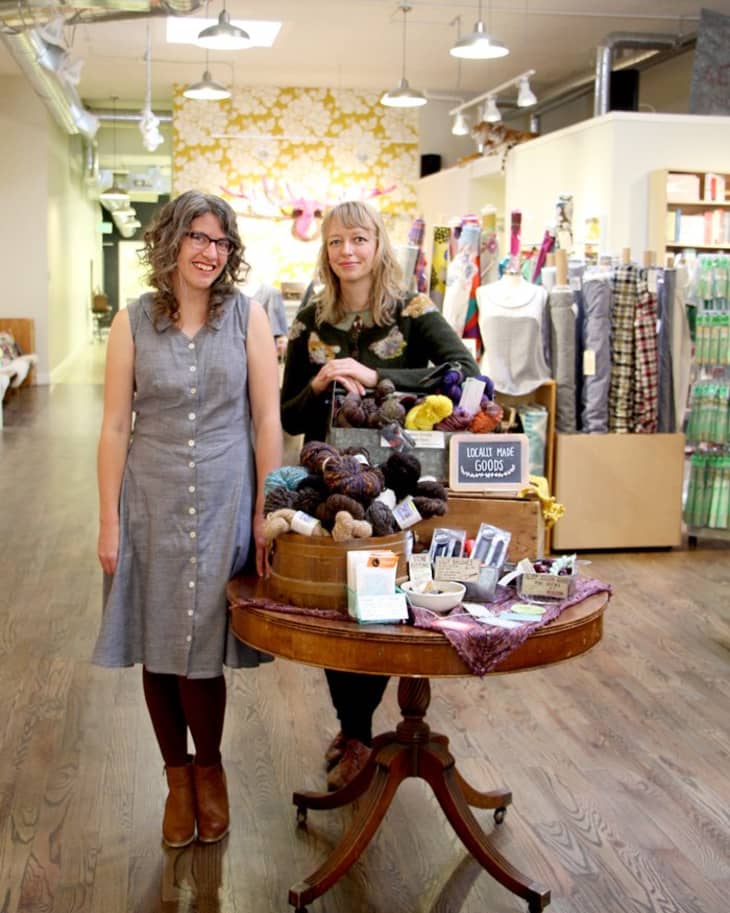 Two women standing in a yarn shop with a table displaying locally made goods and colorful fabrics in the background.