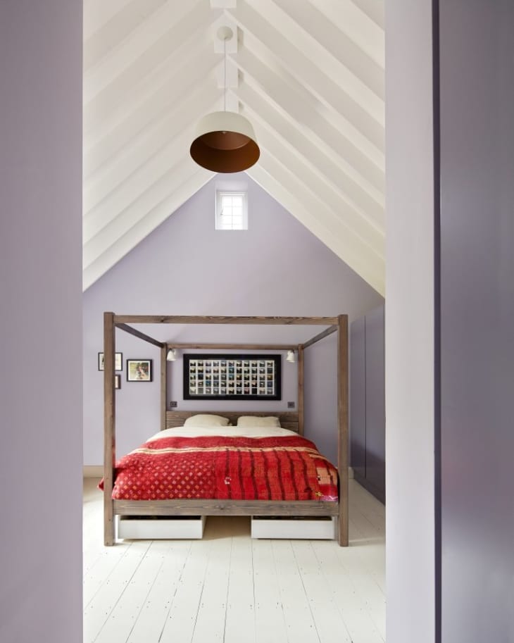Bedroom with vaulted ceiling, wooden canopy bed, red quilt, and framed artwork on lavender walls.