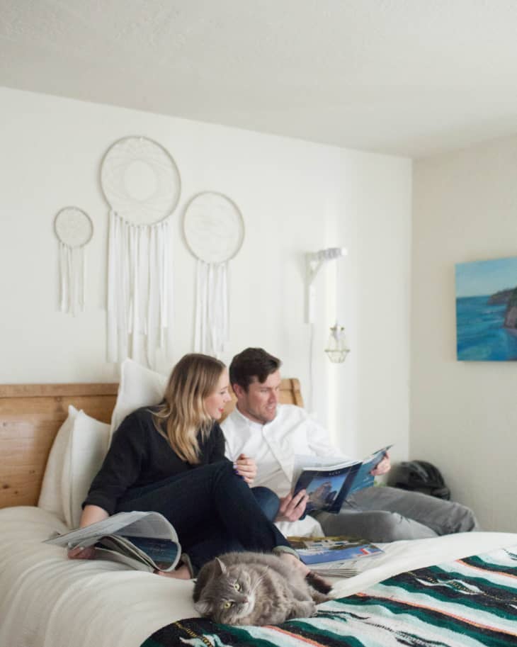 Couple reading magazines on bed with dreamcatchers on wall and a gray cat lying on a colorful blanket.
