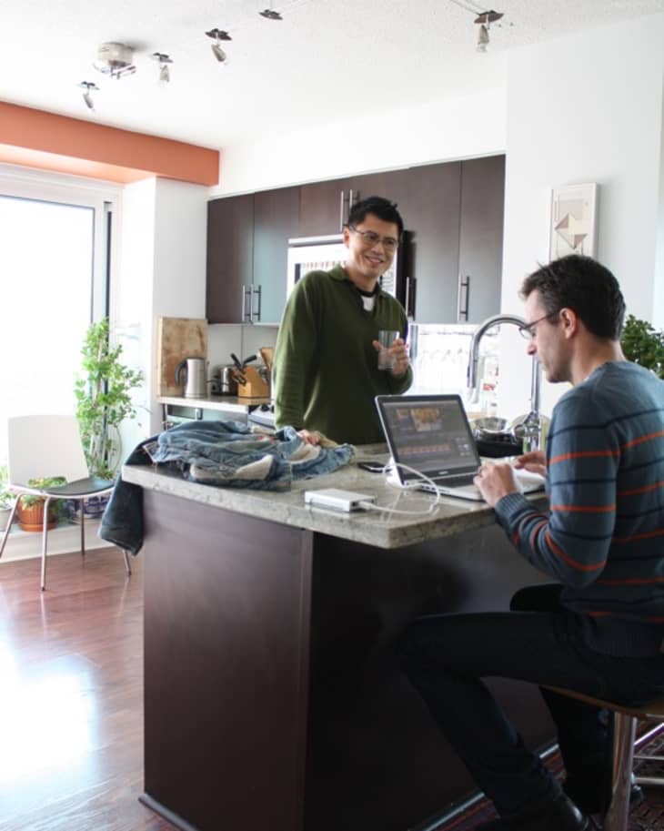 Two men in a modern kitchen, one standing with a drink, the other seated at a laptop on a marble island.