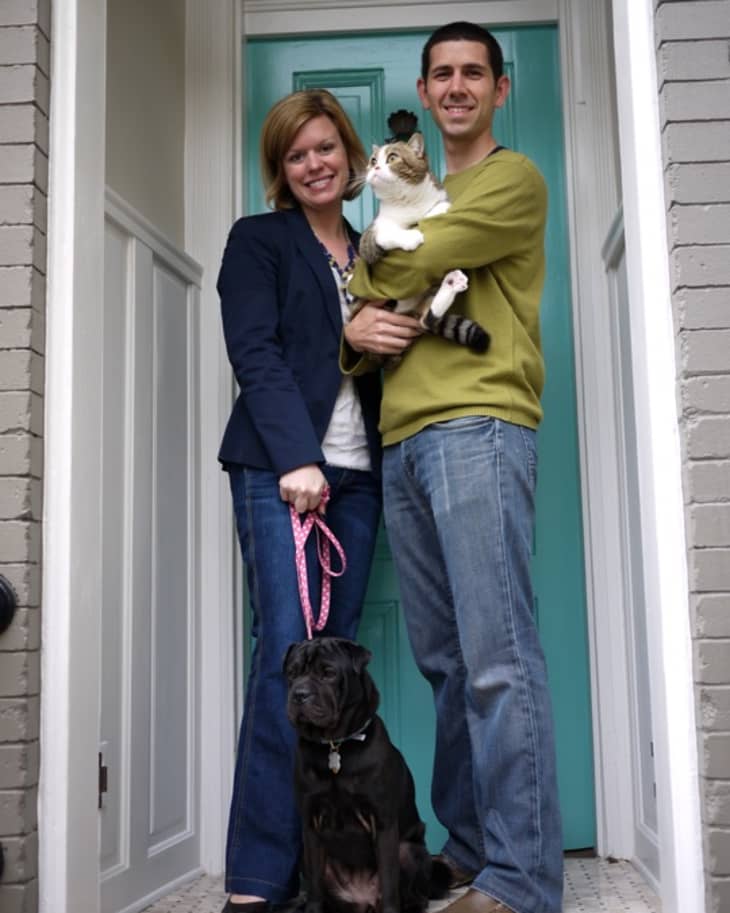 Couple standing in doorway with a cat and a black dog on a leash, teal door in the background.