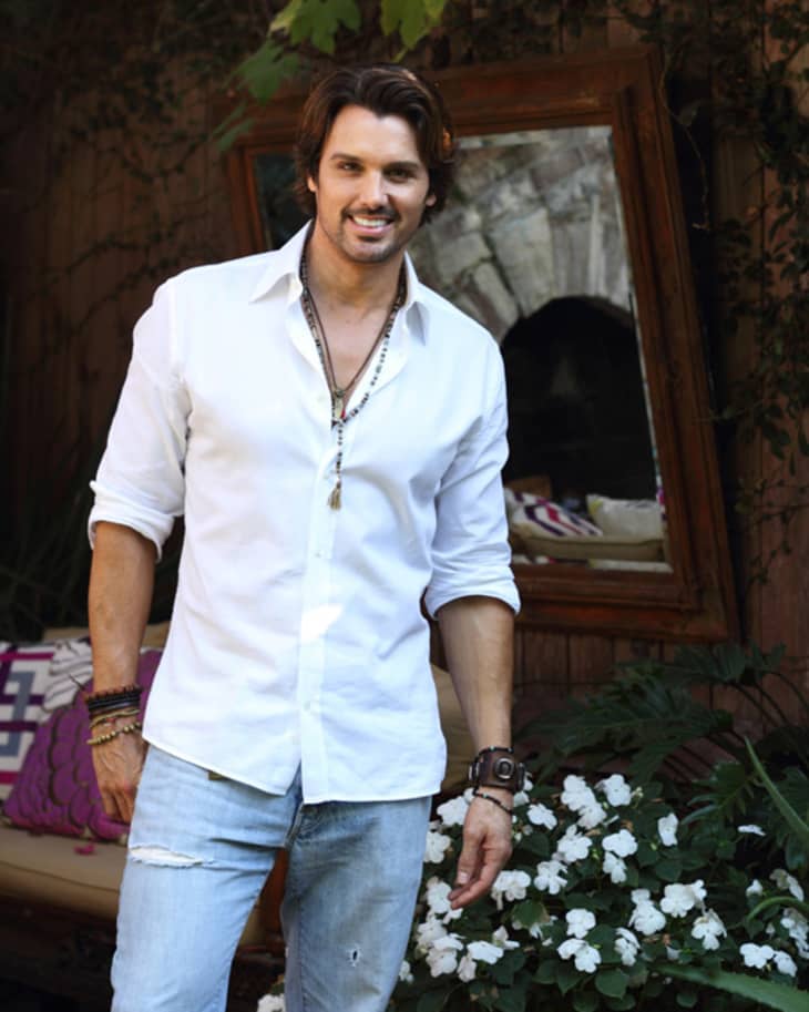 Man in white shirt and jeans standing outdoors near white flowers and a wooden bench with colorful cushions.