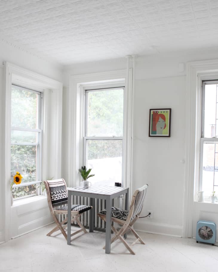 Small dining area with two patterned chairs, a gray table, a sunflower on the windowsill, and a colorful framed artwork.