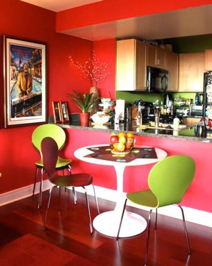 Red dining area with a round white table, green and brown chairs, fruit bowl, and vibrant wall art.