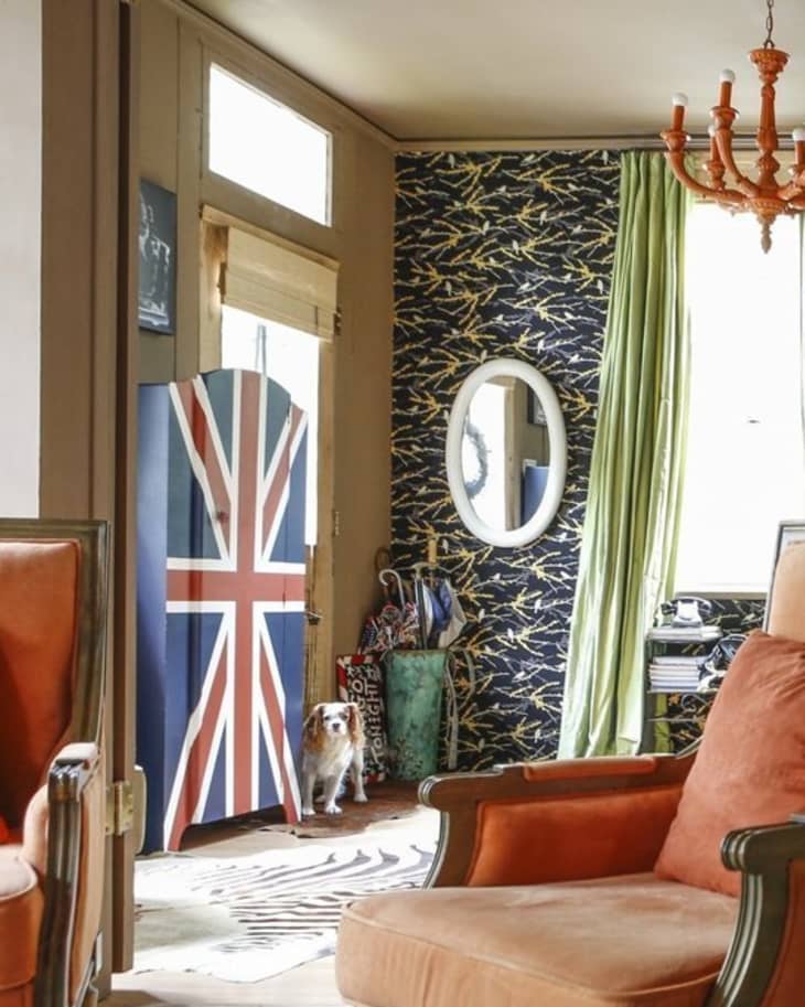 Living room with orange armchairs, Union Jack door, patterned wallpaper, green curtains, and a dog near the entrance.