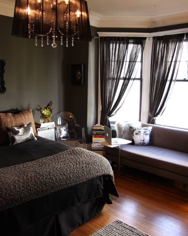 Bedroom with dark walls, a black chandelier, a bed with textured bedding, a sofa with a face pillow, and sheer curtains.