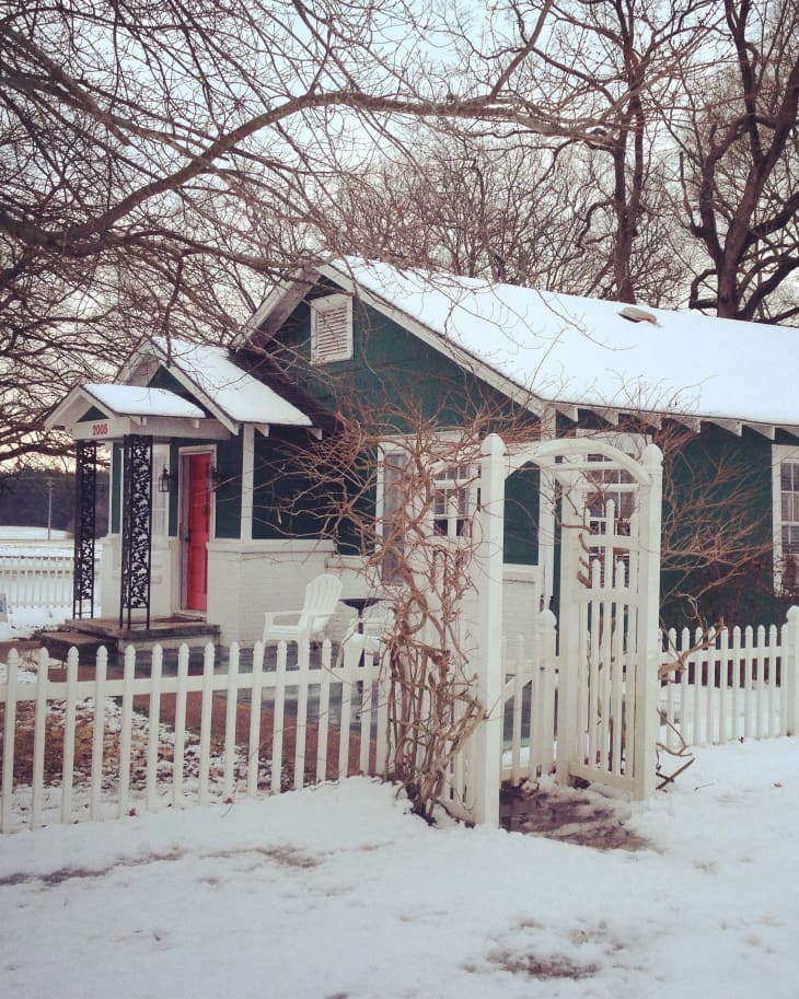 Snow-covered green cottage with red door, white picket fence, and bare trees in winter.