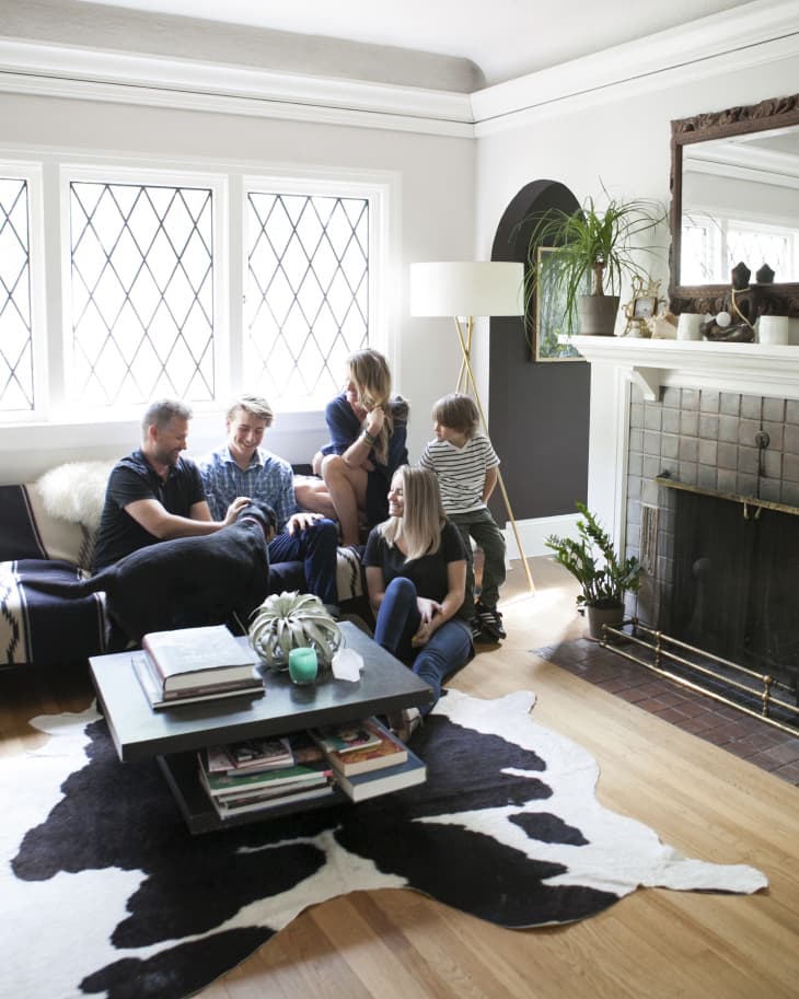 Family sitting on a black sofa in a living room with a cowhide rug, coffee table, and fireplace, surrounded by large windows.