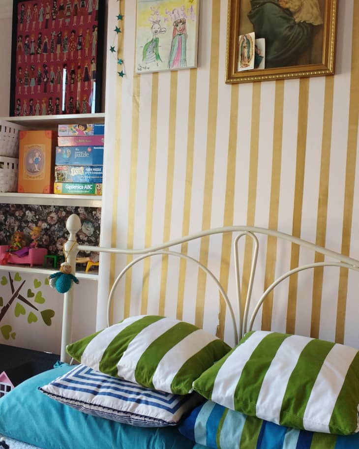 Striped pillows on a bed with a yellow striped wall, framed art, children's drawings, and shelves with board games.