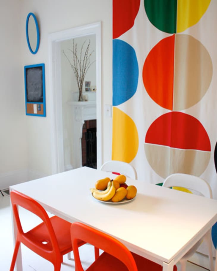 Dining room with white table, red chairs, colorful circle-patterned curtain, and a fruit bowl centerpiece.
