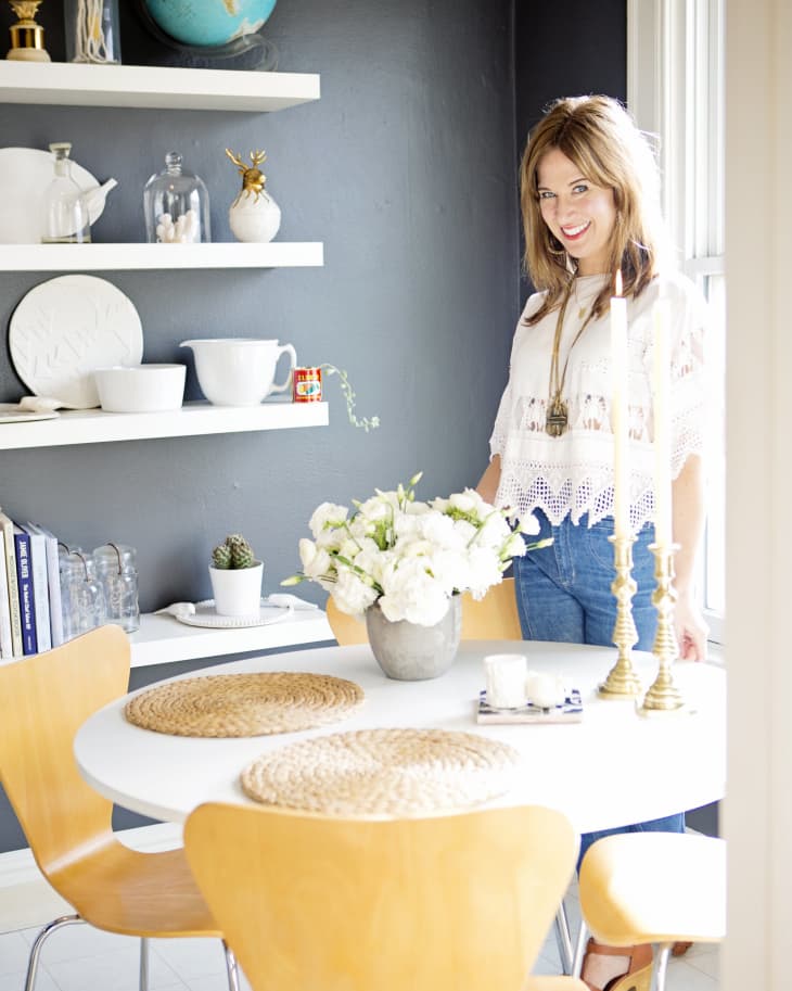 Dining area with wooden chairs, white table, floral centerpiece, and shelves with globe and decor items. Woman standing nearby.