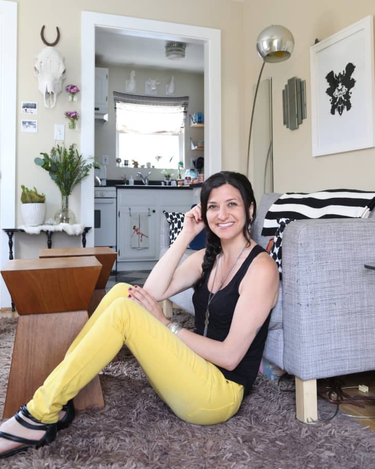 Woman in yellow pants sitting on a shag rug, next to a gray sofa, with a view into a kitchen.