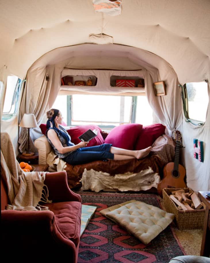 Woman reading on a sofa in a cozy camper with red cushions, guitar, and patterned rug.
