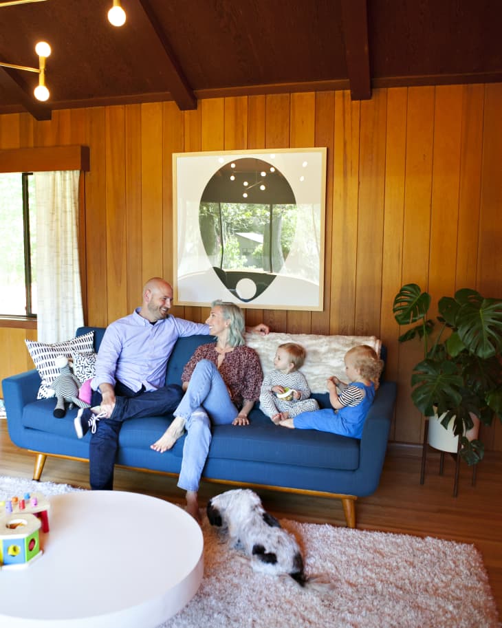 Family sitting on a blue sofa in a wood-paneled living room with a dog, abstract art, and a round white coffee table.