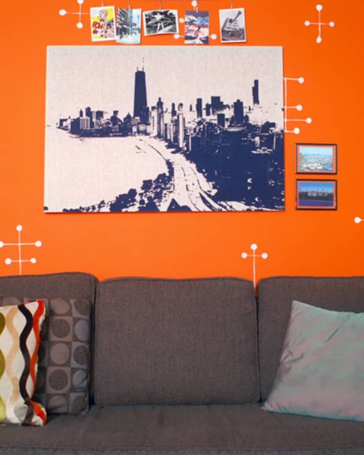 Gray sofa against an orange wall with a cityscape canvas, geometric wall decals, and decorative pillows.