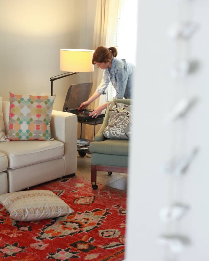 Woman adjusting a record player in a cozy living room with a white sofa, colorful pillows, and a vibrant red patterned rug.