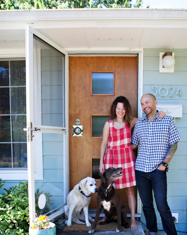 Couple standing with two dogs at the front door of a house, wearing casual summer clothes.