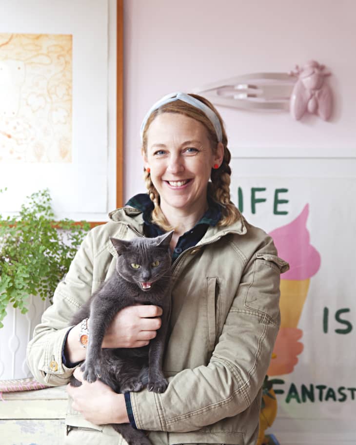 Woman smiling while holding a gray cat, standing in a room with art and plants.