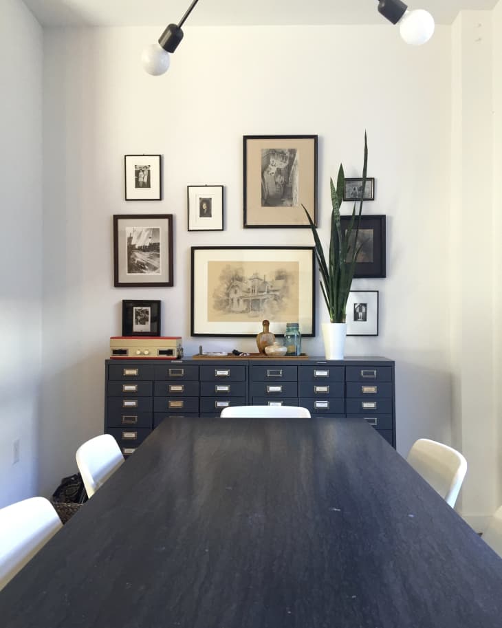 Dining room with dark table, white chairs, black cabinet, framed art, and a tall plant in a white pot.