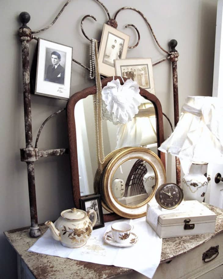 Vintage dresser with ornate mirror, teapot set, framed photos, pearl necklace, and a small clock.