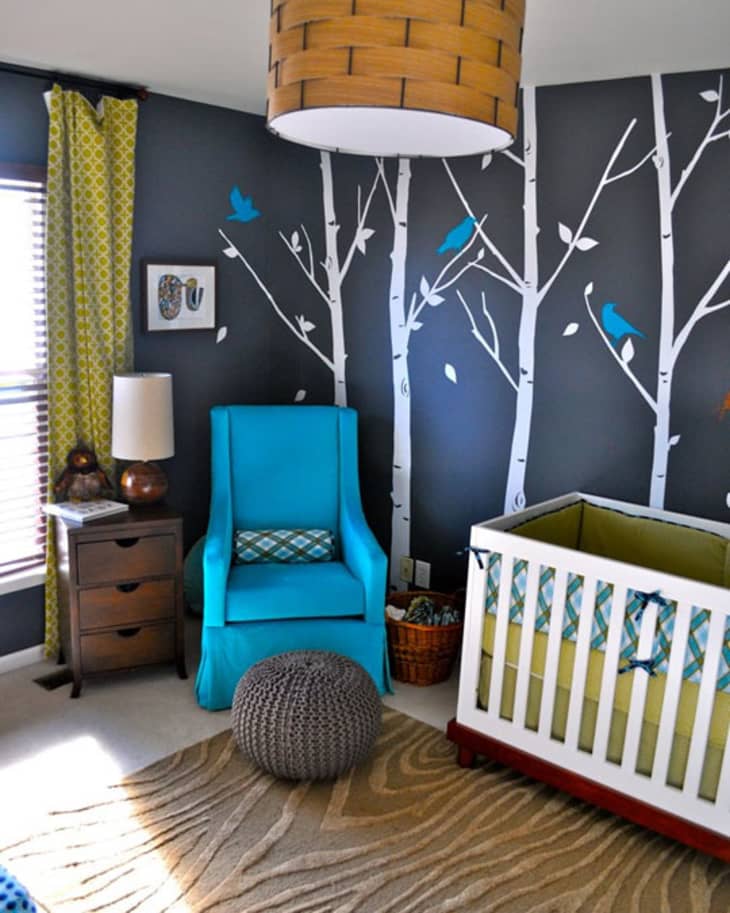 Nursery with tree wall decals, blue chair, white crib, and green curtains.