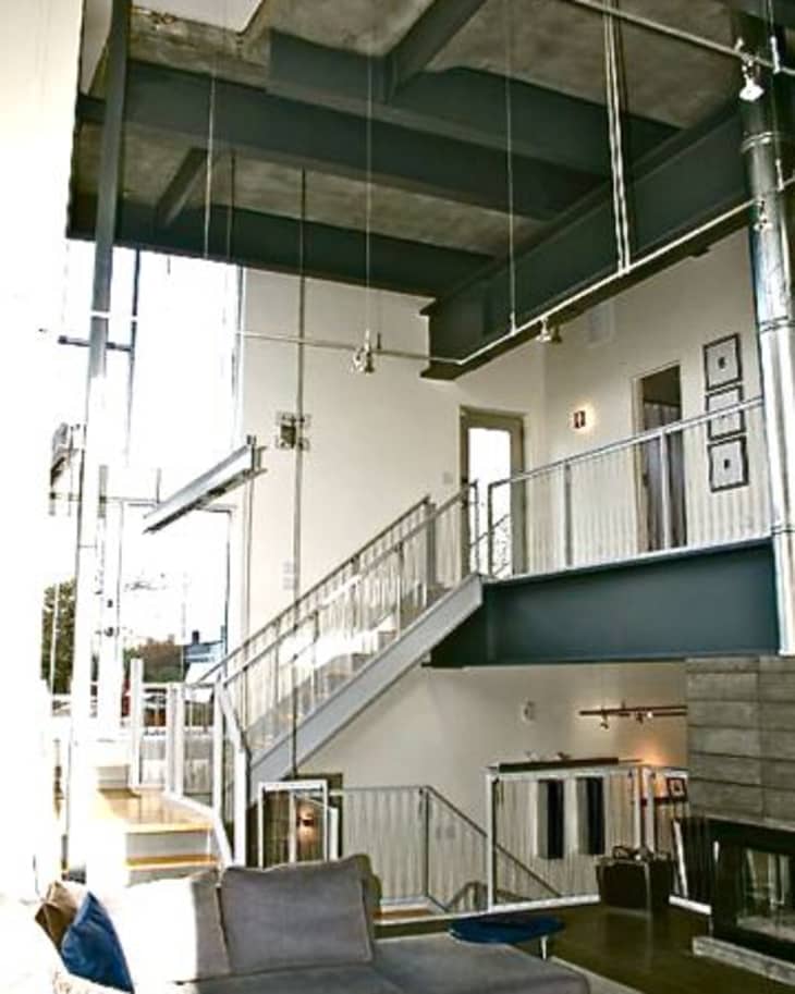 Modern loft with high ceilings, exposed beams, metal staircase, and gray sectional sofa with orange throw blanket.