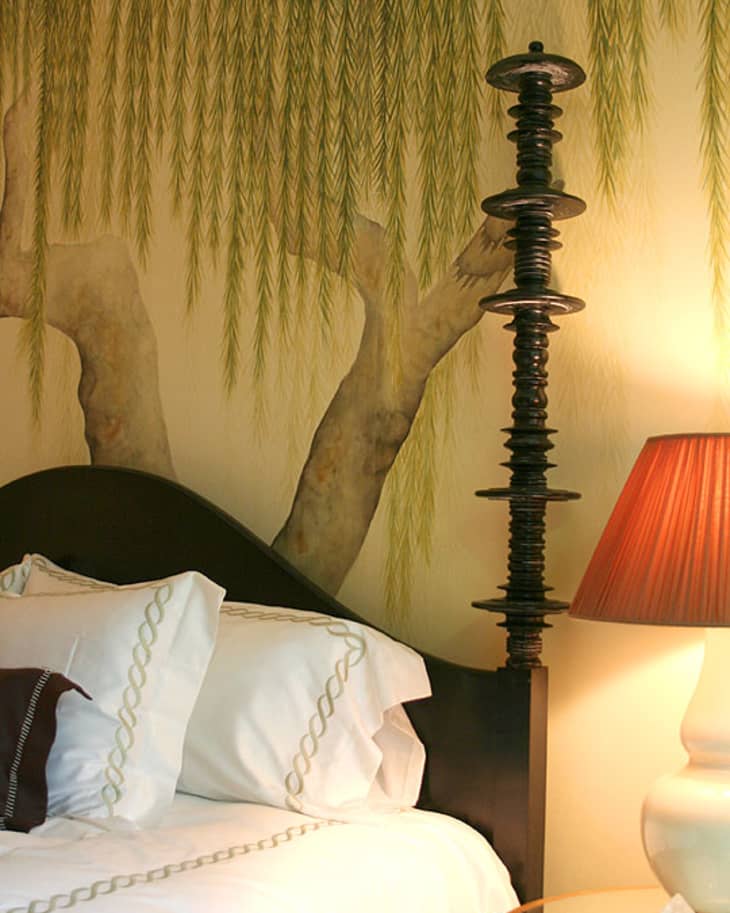 Bed with white linens, decorative pillows, and a unique lamp beside a wall mural of tree branches and hanging leaves.