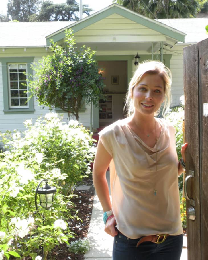 Woman standing at a garden gate in front of a white house with green trim and lush greenery.