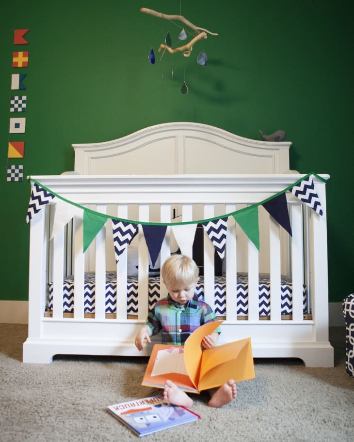 Child reading a book on the floor in front of a white crib with chevron bedding and colorful bunting, green wall background.