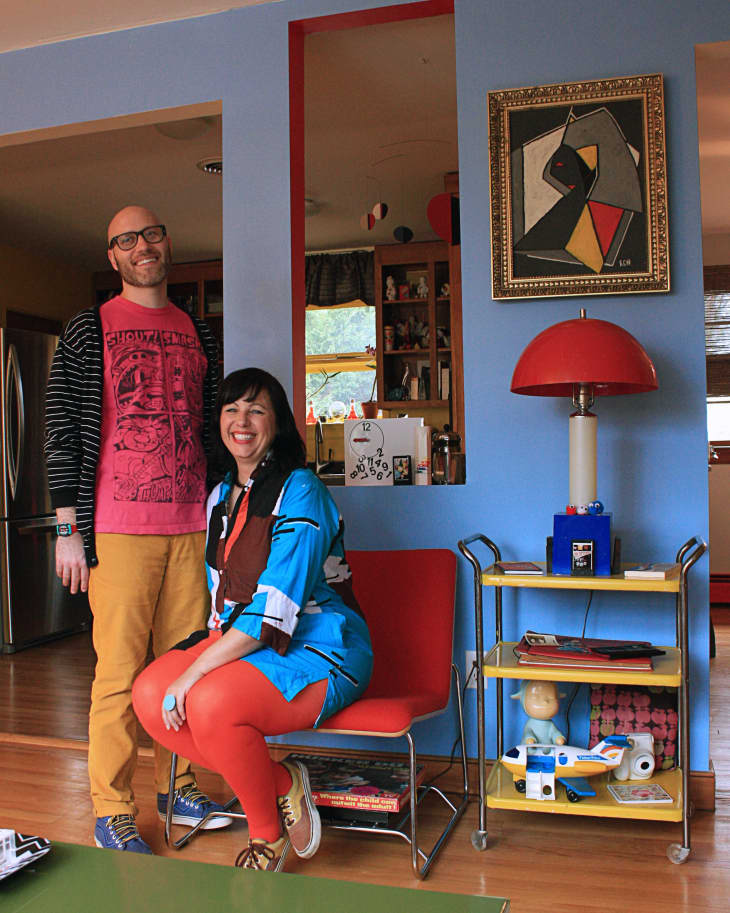 Man and woman in colorful outfits in a living room with blue walls, red chair, and eclectic decor.