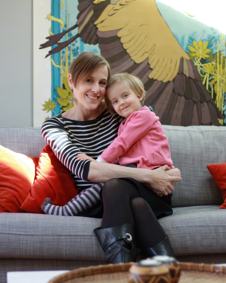 Woman and child sitting on a gray sofa with red cushions, large bird mural in the background.