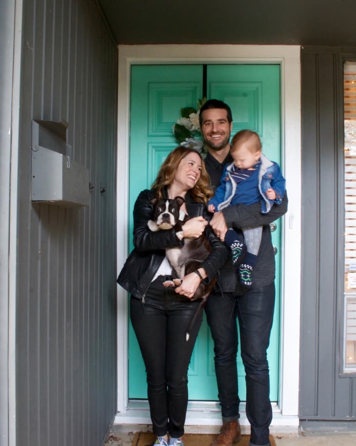 Couple with a baby and a dog standing in front of a turquoise door, smiling.