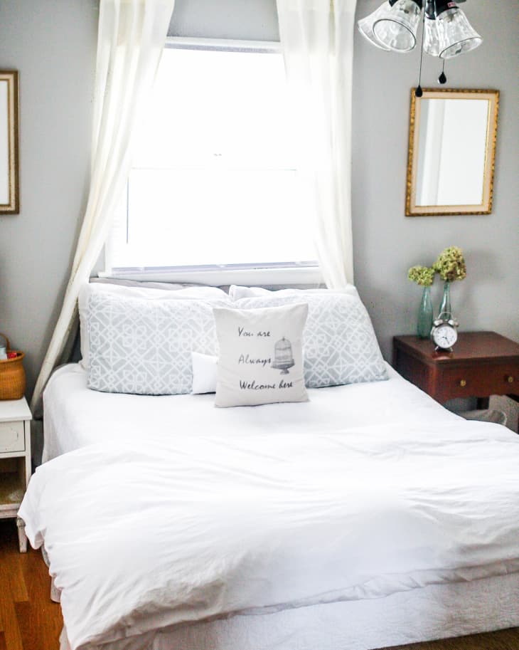 Cozy bedroom with white bedding, decorative pillows, wicker bench, and a ceiling fan, featuring a window with sheer curtains.