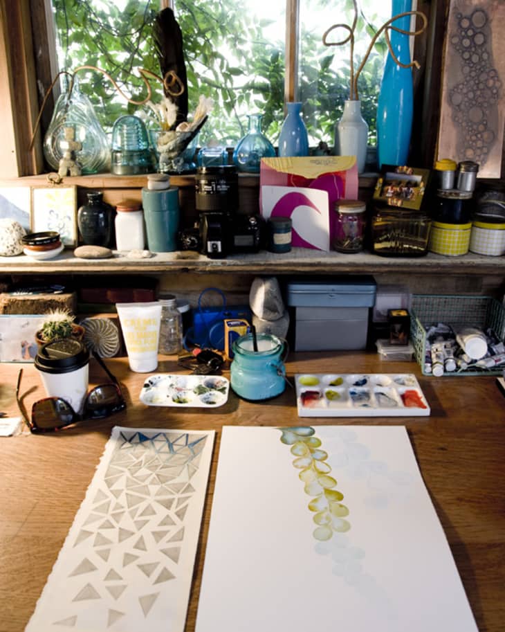 Art studio desk with watercolor paintings, paint palettes, jars, and decorative items on a wooden shelf by a window.